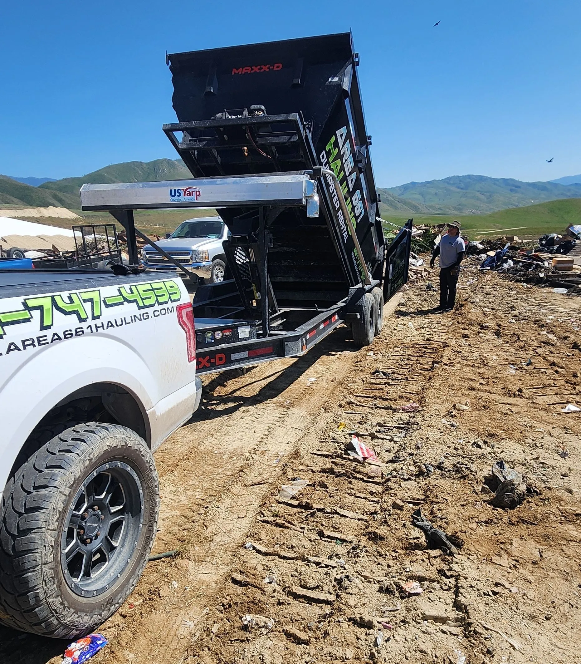 A truck with a dump trailer raised to unload onto dirt ground in an open field, with a person standing nearby in Bakersfield, CA